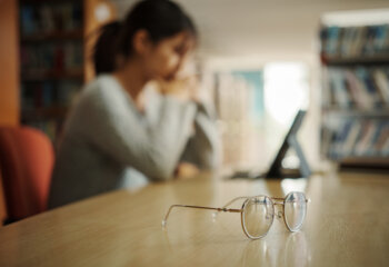 Close up of eyeglasses on table with unhappy girl student using tablet in university library.