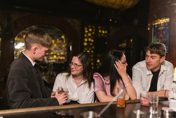 A group of young friends sit at a dimly lit bar, talking and drinking.