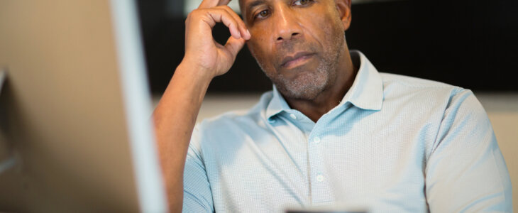 A concerned bald black man with a beard sits at a desk, looking at a computer monitor.