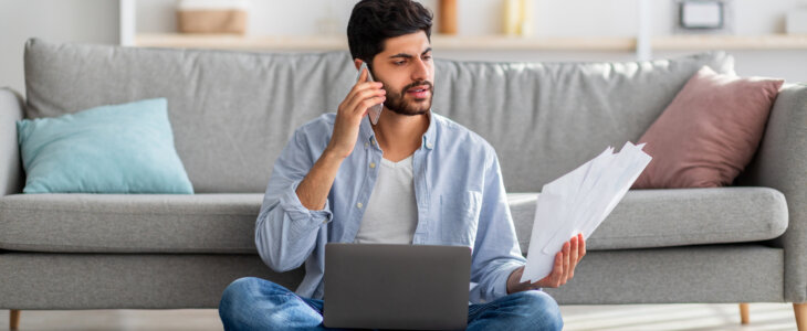 A focused man sitting on the floor with a laptop and papers, talking on a smartphone.