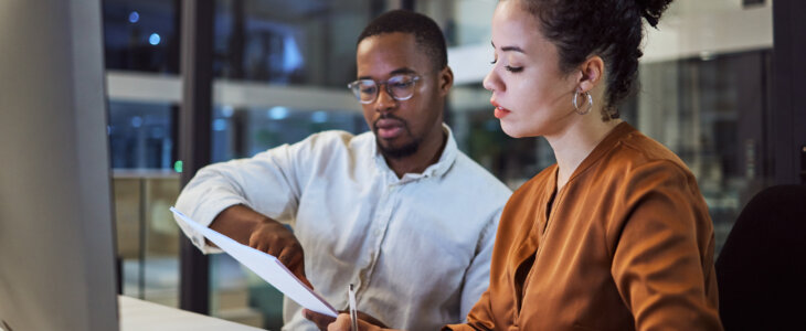 A focused man and woman work together at a desk, with the man pointing at a document.