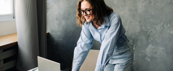 A happy young woman in glasses and a button-up shirt leans over a desk to use a laptop.