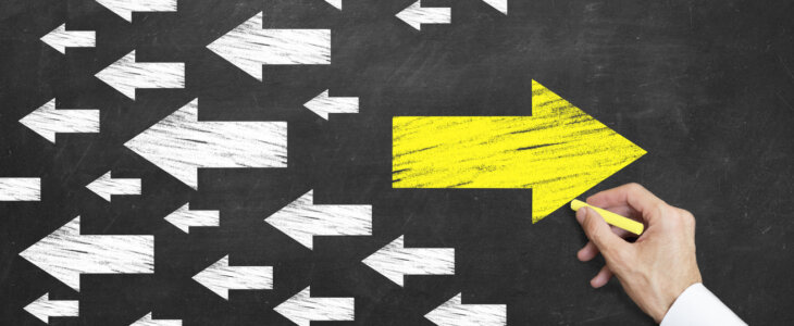 A person drawing a large yellow arrow on a chalkboard, going against a sea of white arrows.