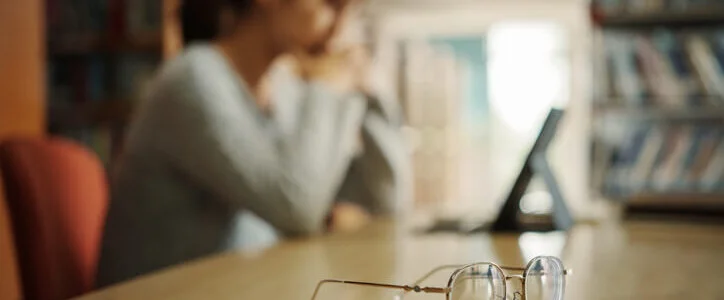 Close up of eyeglasses on table with unhappy girl student using tablet in university library.