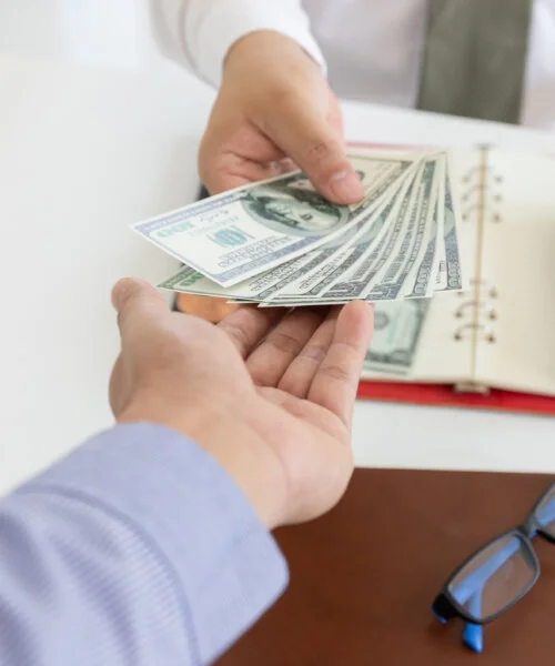 A hand giving another person a stack of one hundred dollar bills.