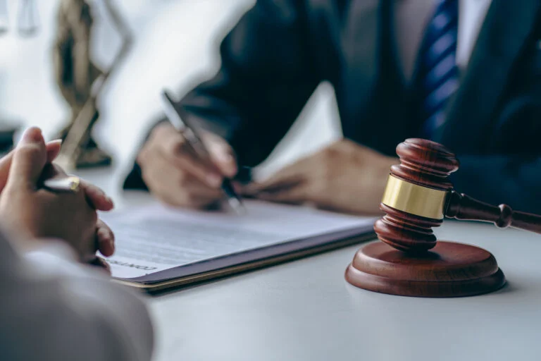 A person signs a legal document with a gavel in the foreground, suggesting a court settlement.