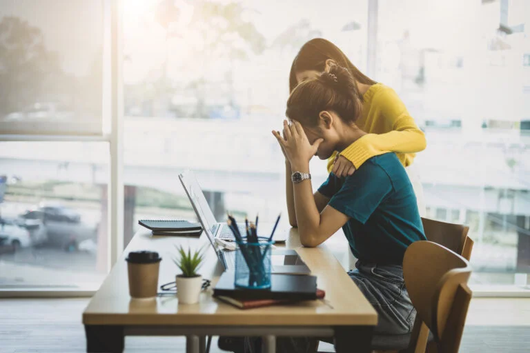 A colleague comforting a stressed coworker who is sitting at her desk with a laptop.