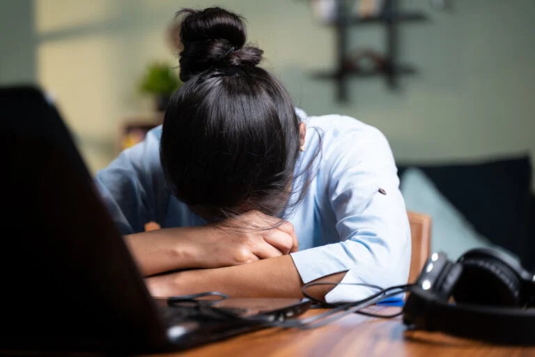 A tired woman with her head down on a desk, with a laptop and headphones nearby.